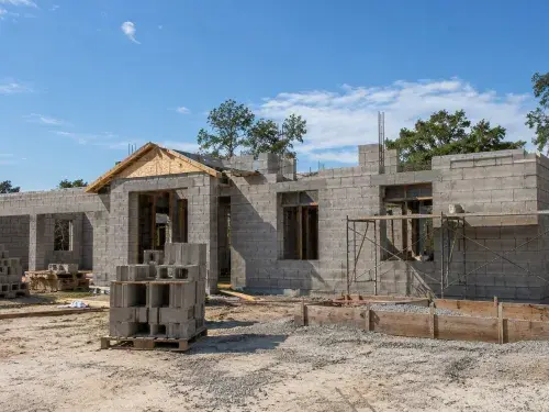 Concrete block house under construction with CMU walls, rebar, scaffolding, and jobsite materials.