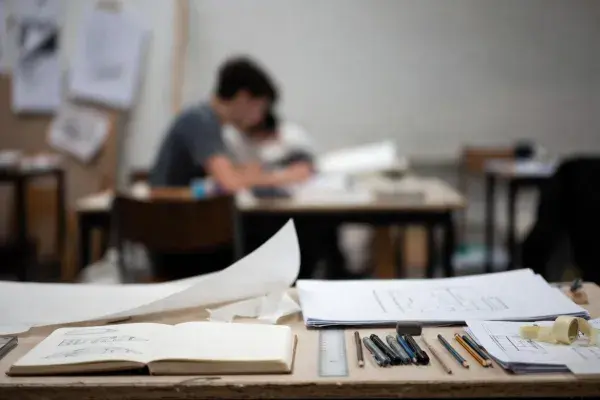 Architectural drawing tools, sketchbook, and plan sheets on a studio desk with students working in the background.