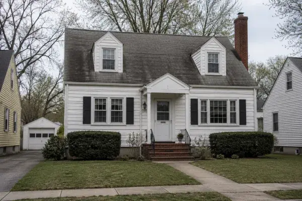 1940s Cape Cod-style house with white siding, dormers, brick chimney, and modest front entry.
