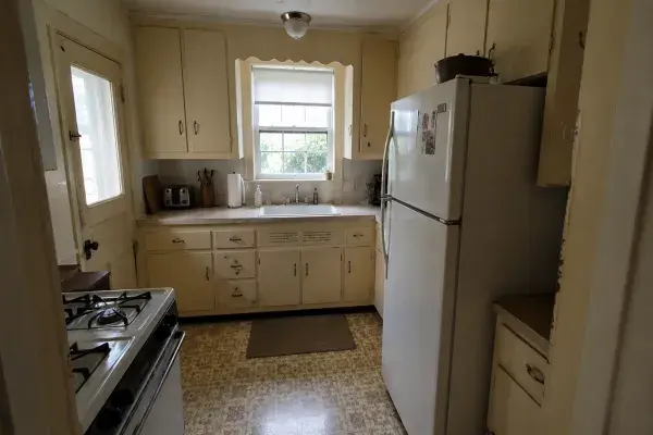 Small 1940s kitchen with older cabinets, limited counter space, dated flooring, weak ceiling light, and a bulky refrigerator crowding the room.