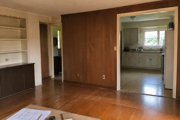 Dated 1960s living room before renovation with wood paneling, worn hardwood floors, an older kitchen opening, and a few planning materials near the edge of the room.