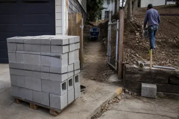 Concrete block pallet staged near a driveway with a narrow access path to the wall location.