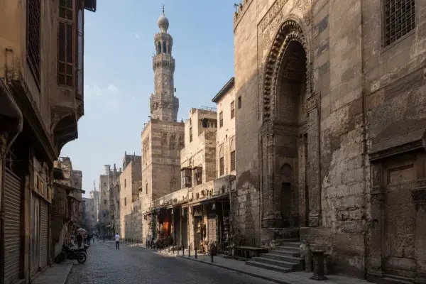 Historic Islamic Cairo street with carved stone portal, minaret, market edge, mashrabiya screens, and narrow shaded buildings.