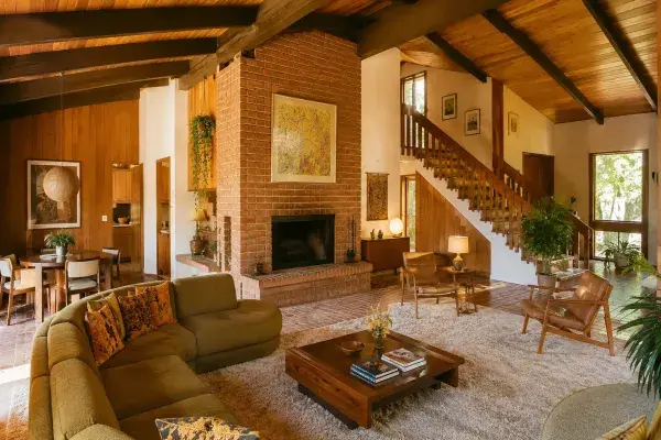Bright 1970s living room with brick fireplace, wood ceiling, open stair, and low seating.