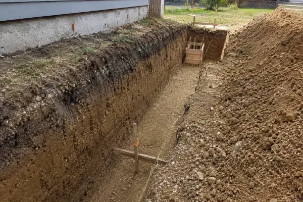 Foundation excavation trench beside a house showing trench depth, soil layers, string lines, and footing layout before concrete placement.